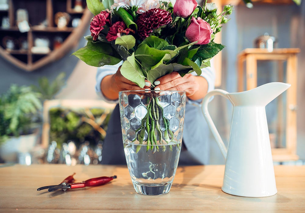 Hands arrange a bouquet of flowers in a hexagonal glass vase filled with water, placed on a wooden table beside pruning shears and a white pitcher, with a blurred indoor background.