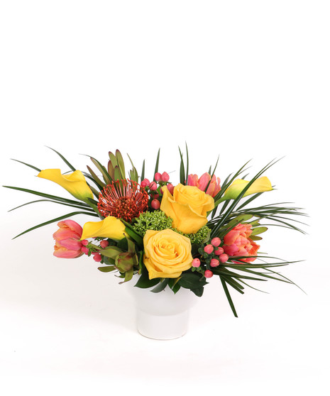 Island Radiance floral arrangement with yellow roses, calla lilies, pincushion proteas, and peach tulips in a white ceramic vase