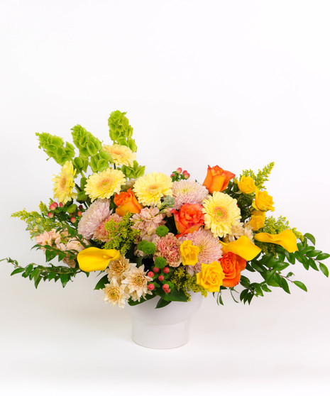 Sunshine & Sage floral arrangement with yellow roses, calla lilies, gerbera daisies, and bells of Ireland in a white ceramic vase