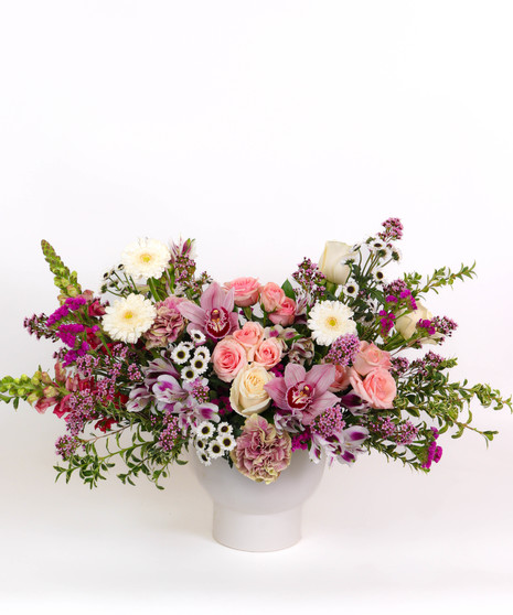 Mauve Melody floral arrangement with mauve roses, orchids, white gerbera daisies, and chamomile in a white ceramic vase